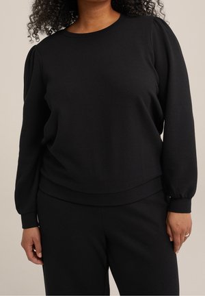 Kobieta ubrana w czarną, teksturowaną bluzkę z długim rękawem i bufkami na ramionach oraz dopasowane czarne spodnie, stojąca na tle jednolitego beżowego tła.