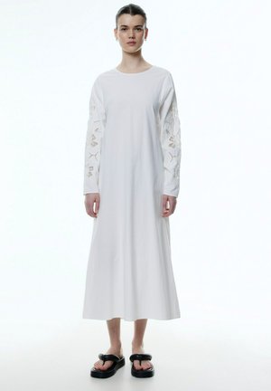 Lange witte jurk met lange mouwen en ingewikkelde kantdetails. De stof is soepel en de silhouet is losvallend. Gemaakt in combinatie met zwarte sandalen.