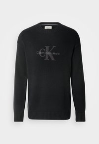 Juodas megztinis su raukšlėta tekstūra, turintis apvalų kaklą ir ilgomis rankovėmis. Parodo pilką siuvinėtą "CK" logotipą su "Calvin Klein Jeans" po juo.