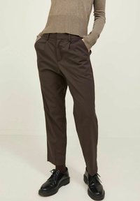 Mörkbruna skräddarsydda byxor med avslappnad passform, har frontveck och sidofickor, tillsammans med svarta snörskor. En ljusstickad topp syns.