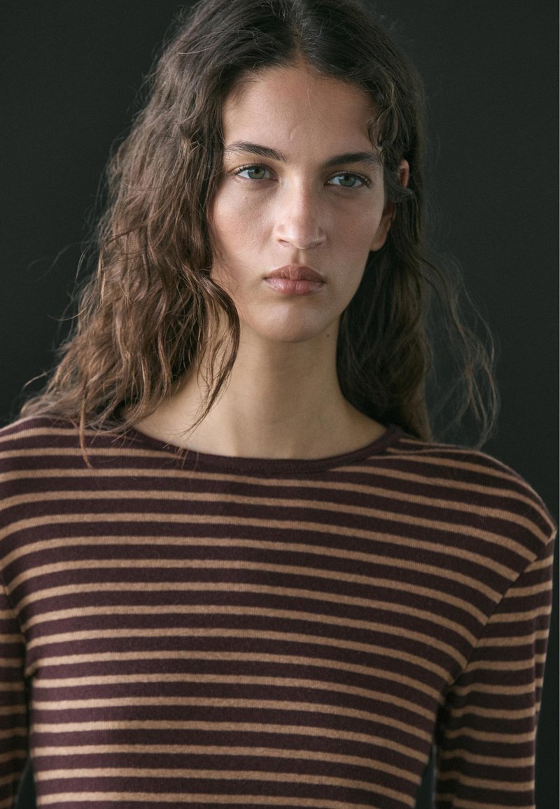 Jeune femme aux cheveux bruns ondulés portant un haut rayé marron et beige, regardant devant elle avec une expression neutre sur un fond sombre.