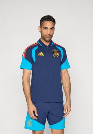 Polo deportivo azul marino con detalles en turquesa, cremallera parcial, logotipo dorado y franjas en rojo, amarillo y azul en los hombros; combinado con pantalones cortos a juego.