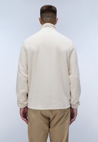 Creme fleece pullover med høj krave, lange ærmer og en blød struktur. Har en glat overflade og et broderet logo nær kanten.