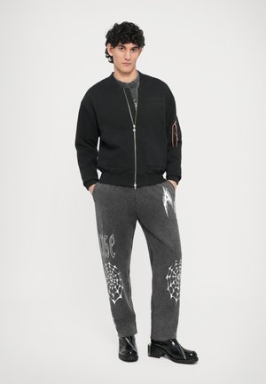 Schwarze Bomberjacke mit Reißverschluss und zwei Vorderentaschen. Graue Jogginghose mit weißen Spinnennetzmustern und grafischem Text. Schwarze glänzende Stiefeletten.