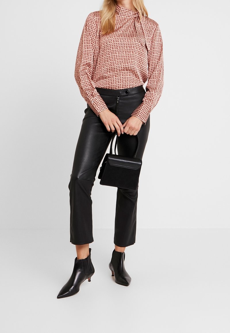 Femme portant un chemisier à manches longues à motifs, un pantalon en cuir noir, des bottines noires et tenant un sac à main noir devant un fond uni.