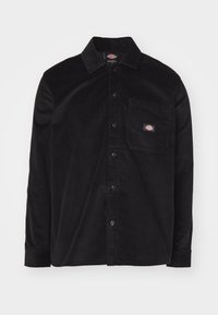 Camisa de pana negra con cuello abotonado, mangas largas, bolsillo frontal y parche con logotipo. Presenta un patrón de tejido texturizado.