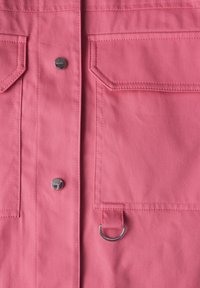 Chemise en tissu rose avec deux poches poitrine et un détail en métal en forme de cercle. Présente des coutures visibles et un bouton-pression métallique brillant.