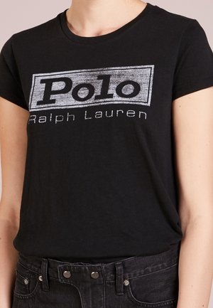 Persoon draagt een zwart T-shirt met een vervaagde witte "Polo Ralph Lauren"-logo en zwarte spijkerbroek.