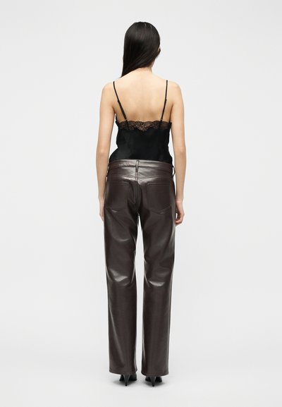 Ženska z dolgimi temnimi lasmi, oblečena v črno camisolo z čipkasto obrobo in temno rjave hlače iz usnja z visokim pasom, stoji z zadnjico obrnjen proti kameri.