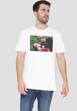 Weißes Baumwoll-T-Shirt mit einem gerahmten Grafikdruck von zwei Figuren, eine in einem roten Outfit, und dem Text "Wie du wünschst" auf einem grünen Hintergrund.