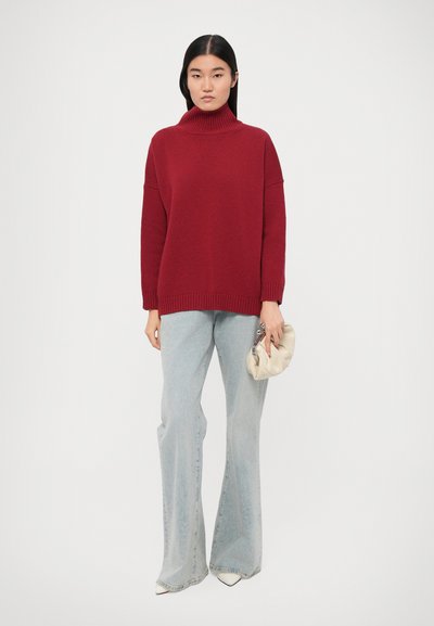 Pull à col roulé rouge en tricot avec coupe décontractée, associé à un jean flare bleu clair. Le mannequin tient une pochette crème.