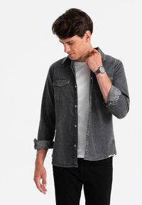 Grijze denimshirting met een knoopsluiting aan de voorkant, twee borstzakken en opgerolde mouwen. Gecombineerd met een grijs t-shirt en zwarte broek.