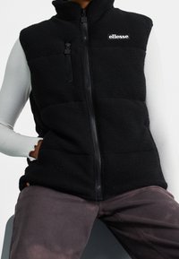 Persona che indossa un gilet in pile nero con logo ellesse, una maglietta a maniche lunghe bianca a coste e pantaloni scuri sbiaditi, mentre è seduta.