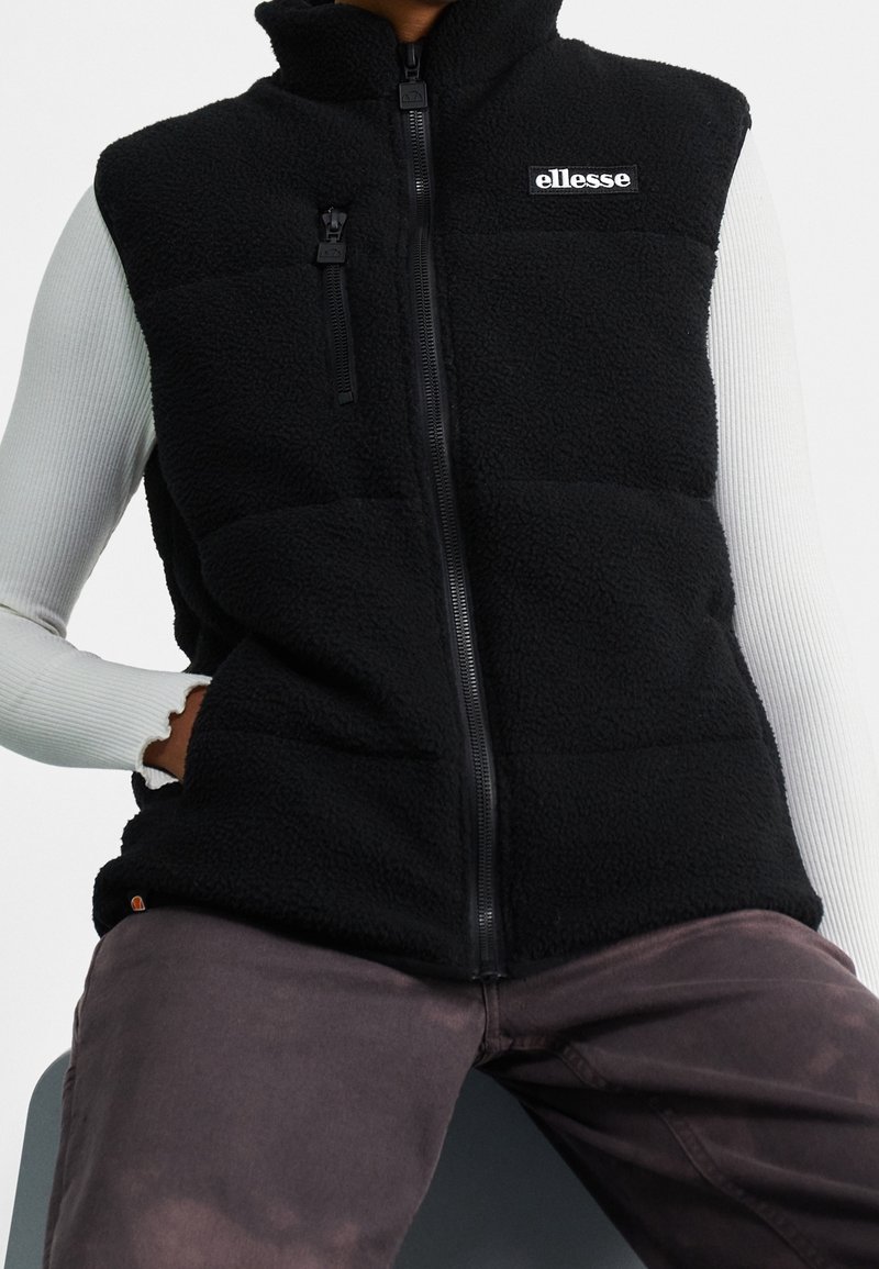 Persona che indossa un gilet in pile nero con logo ellesse, una maglietta a maniche lunghe bianca a coste e pantaloni scuri sbiaditi, mentre è seduta.