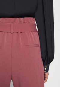 Roze hooggesneden broek met een gegatherde tailleband en twee zakken aan de voorkant, gecombineerd met een zwarte blouse met lange mouwen en gouden knoopaccenten.