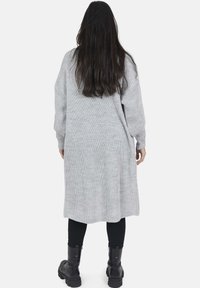 Lungo cardigan grigio lavorato a maglia con spacco laterale, texture a coste e maniche oversize. Indossato sopra leggings neri e stivali neri chunky.