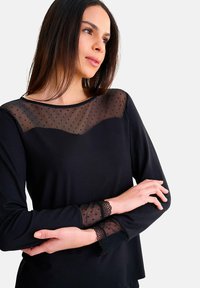 Haut noir avec une superposition en organza à pois à l'encolure et aux manches, ornée de dentelle. Manches longues et design décontracté.