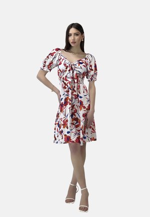 Blumenkleid in Weiß mit roten und blauen Blumen. Puffärmel, Bindeband vorne und geraffte Taille. Kombiniert mit weißen Sandalen mit Riemchen.