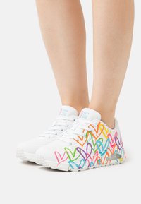 Weiße Sneaker mit einem bunten Herzmuster in Pink, Orange, Blau und Grün. Sie verfügen über eine strukturierte Oberfläche und eine sichtbare Luftpolsterung.