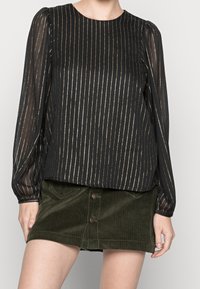 Blusa nera a maniche lunghe con righe verticali dorate, maniche a palloncino, scollatura rotonda, abbinata a una gonna in velluto a coste verde scuro con bottoni frontali.