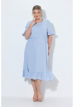 Donna con capelli biondi che indossa un vestito azzurro chiaro di media lunghezza con maniche e orlo arricciati, in piedi davanti a uno sfondo bianco semplice.