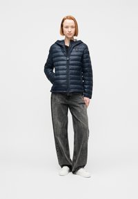 Γυναίκα που στέκεται με τα χέρια στις τσέπες, φοράει ένα μπλε σκούρο μπουφάν τύπου πάπλωμα, άνετο γκρι τζιν και λευκά αθλητικά παπούτσια σε απλό λευκό φόντο.