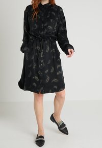 Svart klänning med knäppning fram, långa ärmar och dragsko i midjan. Har ett subtilt mönster och lättviktig tyg. Bärs med svarta ballerinas.