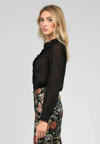 Blouse noire en voile avec des volants, avec un devant à boutons ; assortie à une jupe à motif floral présentant des fleurs colorées sur un fond sombre.