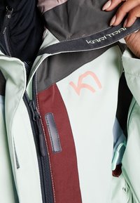 Jasnozielona kurtka z bordowym akcentem, szarym kołnierzem, wieloma zamkami błyskawicznymi i wyraźnym logo. Gładka tekstura z widocznymi detalami przeszyć.