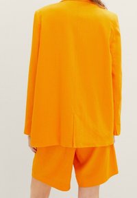 Oranžový lněný blazer s klopou a větráním na zádech, spárovaný s ladícími krajkovými šortkami, ukazující uvolněnou siluetu a hladkou strukturu.