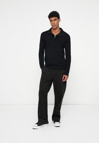 Schwarzes Poloshirt mit langen Ärmeln und einem Viertelreißverschluss, kombiniert mit lockeren schwarzen Hosen und schwarzen Sneakern mit weißen Sohlen. Minimalistisches Design.