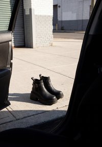 Bottines noires en cuir avec boucles posées sur un trottoir en béton ensoleillé, vues de l'intérieur d'une portière de voiture ouverte.