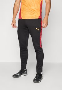 Sorte atletiske bukser med røde sidestriber, med et guld Puma-logo på låret. Lavet af strækbart materiale, tætsiddende pasform. Sorte sportssko.