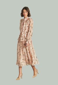 Vestido midi floral en rosa claro con volantes y un cuello atado, con hojas verdes y flores melocotón. Combinado con tacones altos beige.