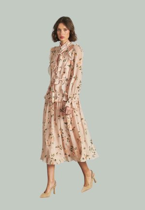 Vestido midi floral en rosa claro con volantes y un cuello atado, con hojas verdes y flores melocotón. Combinado con tacones altos beige.