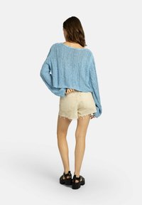Světlomodrý pletený krátký svetr s volným střihem, kombinovaný s opranými béžovými šortkami a černými sandály s otevřenou špičkou. Textura působí vzdušně a uvolněně.