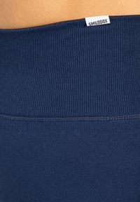 Marineblauwe ribstof tailleband met een klein wit label waarop "SMILODOX" staat. Gladde textuur met zichtbare naden aan de basis.