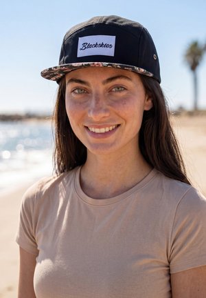 Noor naine, kes kannab musta "Blackskies" mütsi ja beežikat särki, naeratab päikeselisel rannal, kus taustal on uduse veega ja palmi puu.