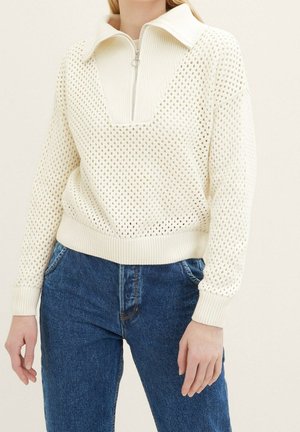 Pull en tricot couleur crème avec un demi-zip, un décolleté en V et un motif en losanges ; accompagné d'un jean en denim bleu foncé.