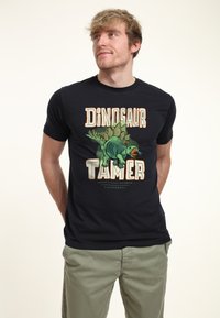 Joven vistiendo una camiseta negra con una ilustración de estegosaurio verde y el texto "Domador de Dinosaurios California", de pie con las manos detrás de la espalda.