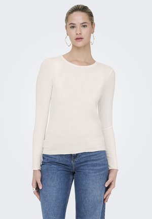 Joven con cabello rubio recogido, lleva grandes pendientes de aro, una camiseta blanca ajustada de manga larga y vaqueros azules, sobre un fondo liso.