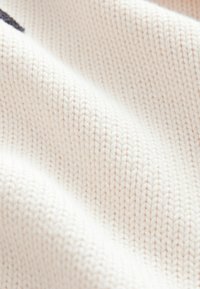 Tissu tricoté texturé, beige clair, avec des points fins et réguliers, créant une surface douce. Aucun motif ou embellissement distinct visible.