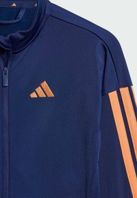 Námornícka športová bunda s oranžovým trojitým pruhom. Obsahuje vysoký golier, plný zips a textilný materiál s logom značky na hrudi.