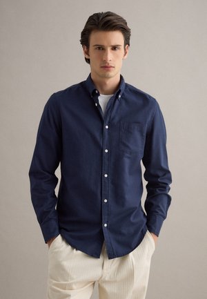 Junger Mann steht mit Händen in den Taschen, trägt ein navyfarbenes Hemd über einem weißen T-Shirt und beige Hosen vor einem neutralen Hintergrund.