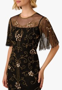 Žena v černých šatech s průhlednými sí ťovanými rukávy a zlatou květinovou výšivkou z flitrů, stojící s jednou rukou uvolněnou podél těla.