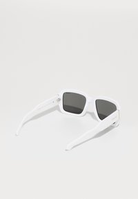 Lunettes de soleil blanches en plastique avec des verres carrés, des verres teintés gris, des accents en métal et le logo de la marque sur une branche.