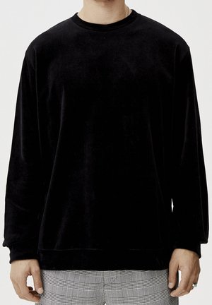 Personne portant une chemise en velours noire à manches longues avec col rond et un pantalon à carreaux gris, les mains détendues sur les côtés contre un fond clair.