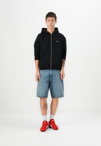 Black zippered hoodie with front pockets, worn over light blue denim shorts. Accessorized with red sneakers and gray socks.