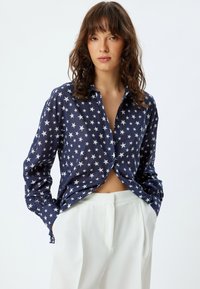Camisa de botones azul marino con un patrón de estrellas blancas, con cuello y mangas largas. Combinada con pantalones blancos de tiro alto.
