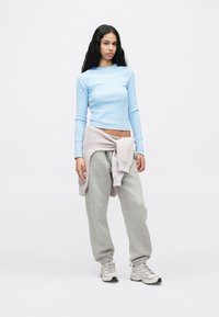 Νεαρή γυναίκα που στέκεται μπροστά σε λευκό φόντο, φορώντας ένα ανοιχτό μπλε μακρυμάνικο μπλουζάκι, γκρι φόρμες, μπεζ αθλητικά παπούτσια και ένα πουλόβερ δεμένο γύρω από τη μέση.